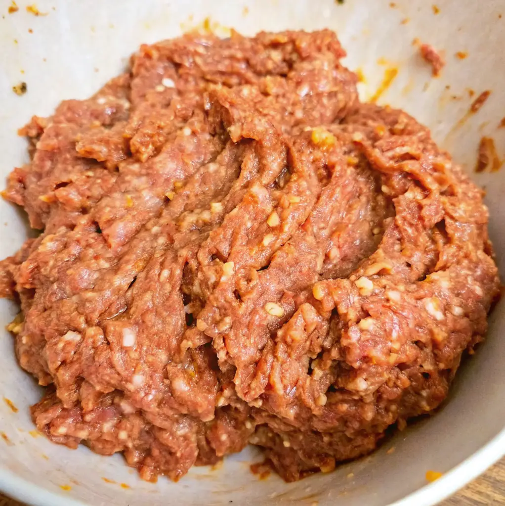 meatloaf mixture after being thoroughly combined, featuring ground beef, visible white pieces of fat or breadcrumbs, and flecks of spice in a white bowl.