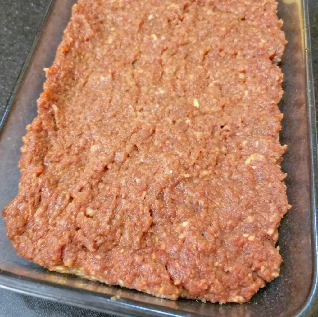 meatloaf mixture pressed into the bottom of a rectangular glass baking dish, forming a flat, even base layer for a casserole.