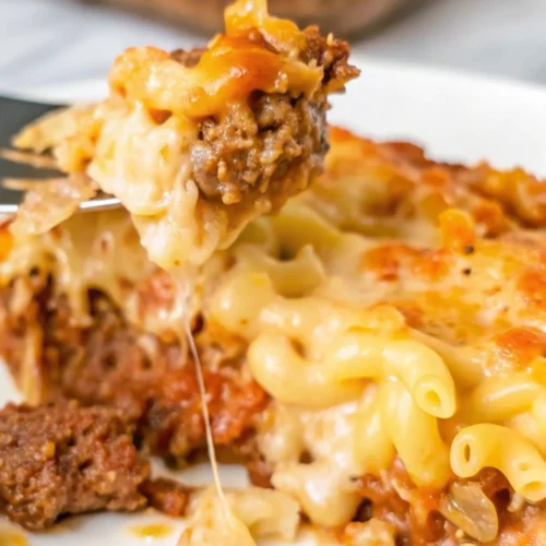 This close-up shot of a serving of mac and cheese meatloaf casserole shows a fork lifting a bite of the cheesy pasta and meatloaf, with strands of melted cheese stretching below.