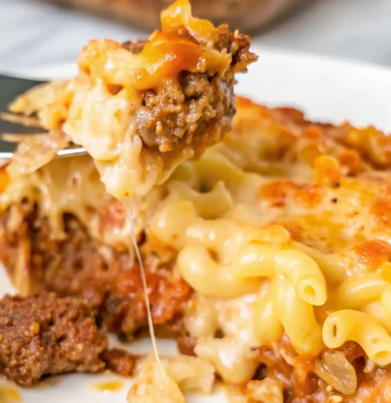 This close-up shot of a serving of mac and cheese meatloaf casserole shows a fork lifting a bite of the cheesy pasta and meatloaf, with strands of melted cheese stretching below.