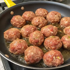 A close-up view of a non-stick skillet on a stovetop, filled with approximately a dozen raw Mississippi Meatballs browning in hot oil.