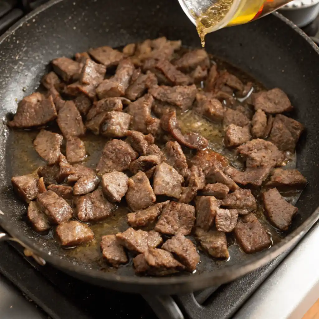 Olive oil being poured into a skillet with seared, golden-brown beef strips on a stovetop.