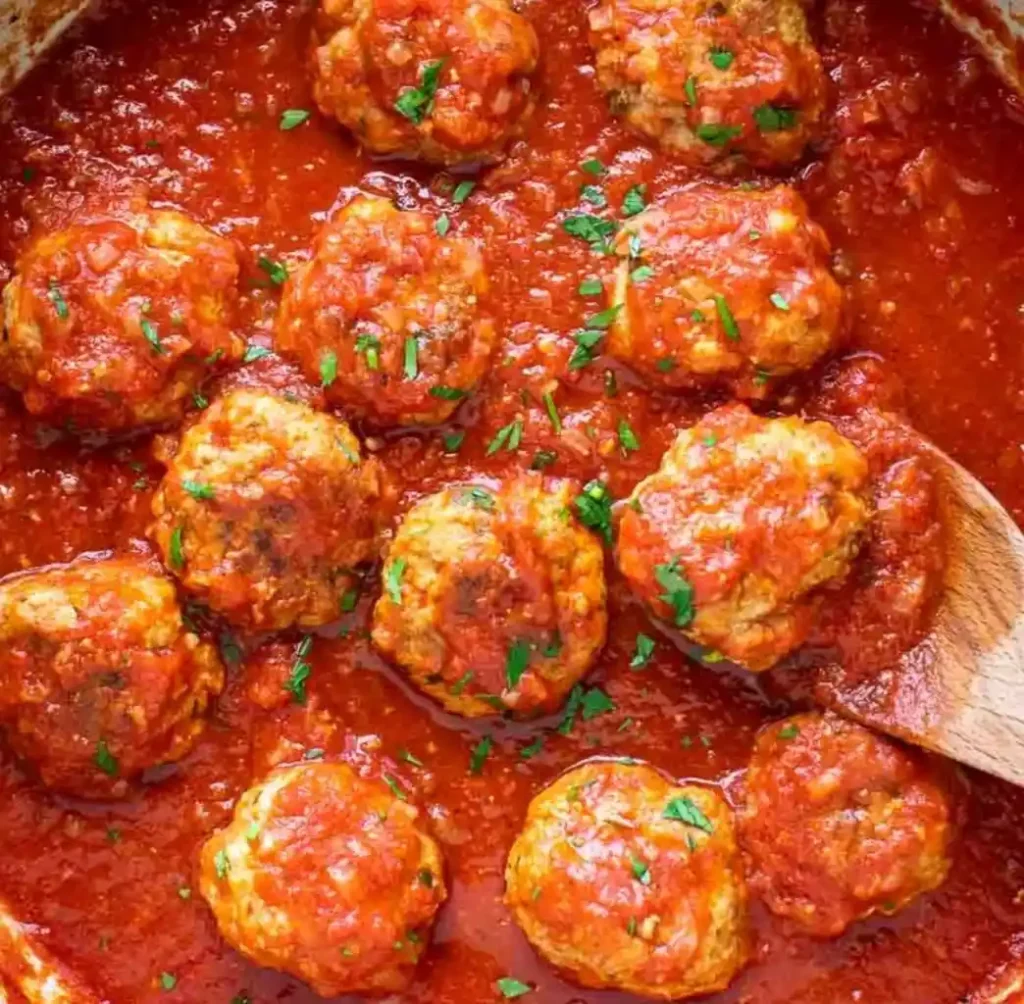 nine large Wagyu meatballs simmering in a rich, bright red tomato sauce speckled with herbs, parsley. A wooden spoon is visible on the right side of the pan.