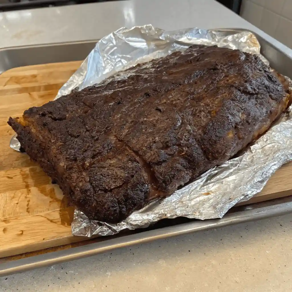 A dark, cooked brisket, still in foil, resting on a wooden board.