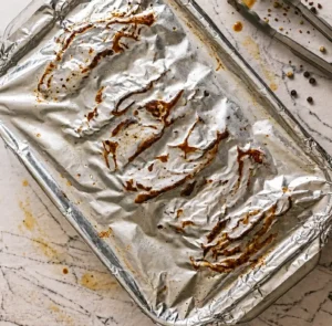 Oven-baked beef country-style ribs tightly wrapped in aluminum foil inside a glass baking dish, resting on a light marble