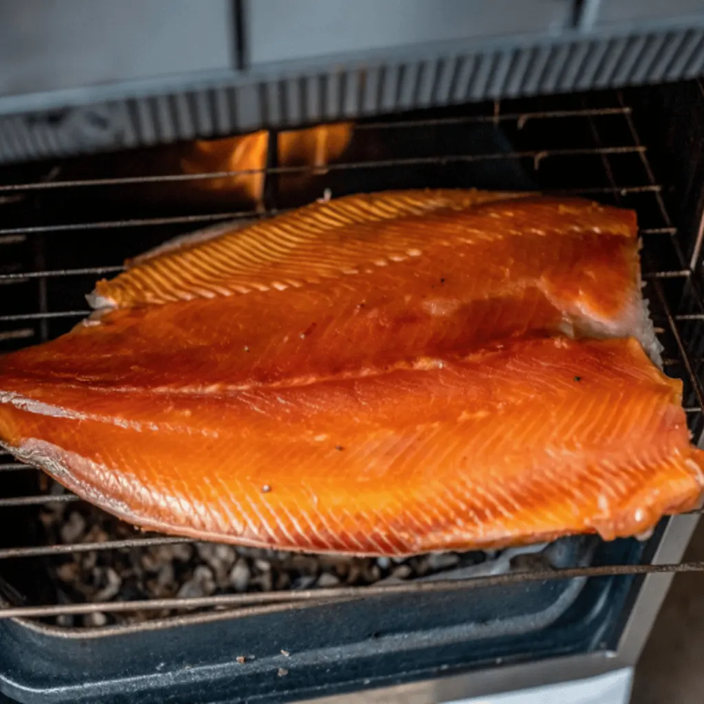 A whole salmon fillet with its skin on is being cold-smoked in a smoker. The vibrant orange fillet rests on a wire rack above smoldering wood chips, with a subtle flame or heat source visible in the background.