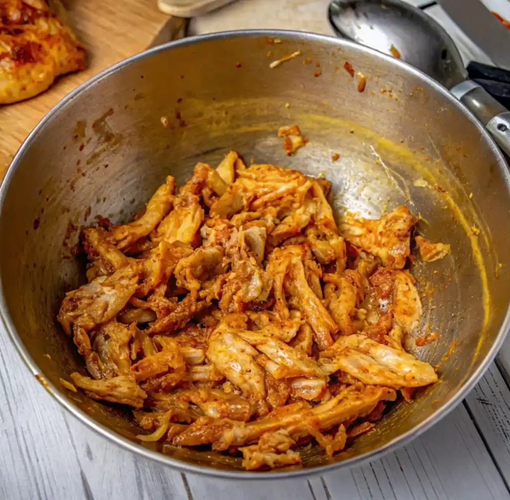 A bowl of brightly seasoned, pulled chicken, fully mixed with a thick birria chili sauce, resting on white painted wood.