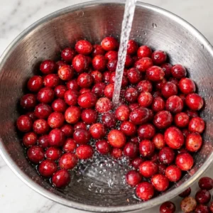 Fresh red cranberries being thoroughly rinsed with running water in a stainless steel bowl on a marble surface.