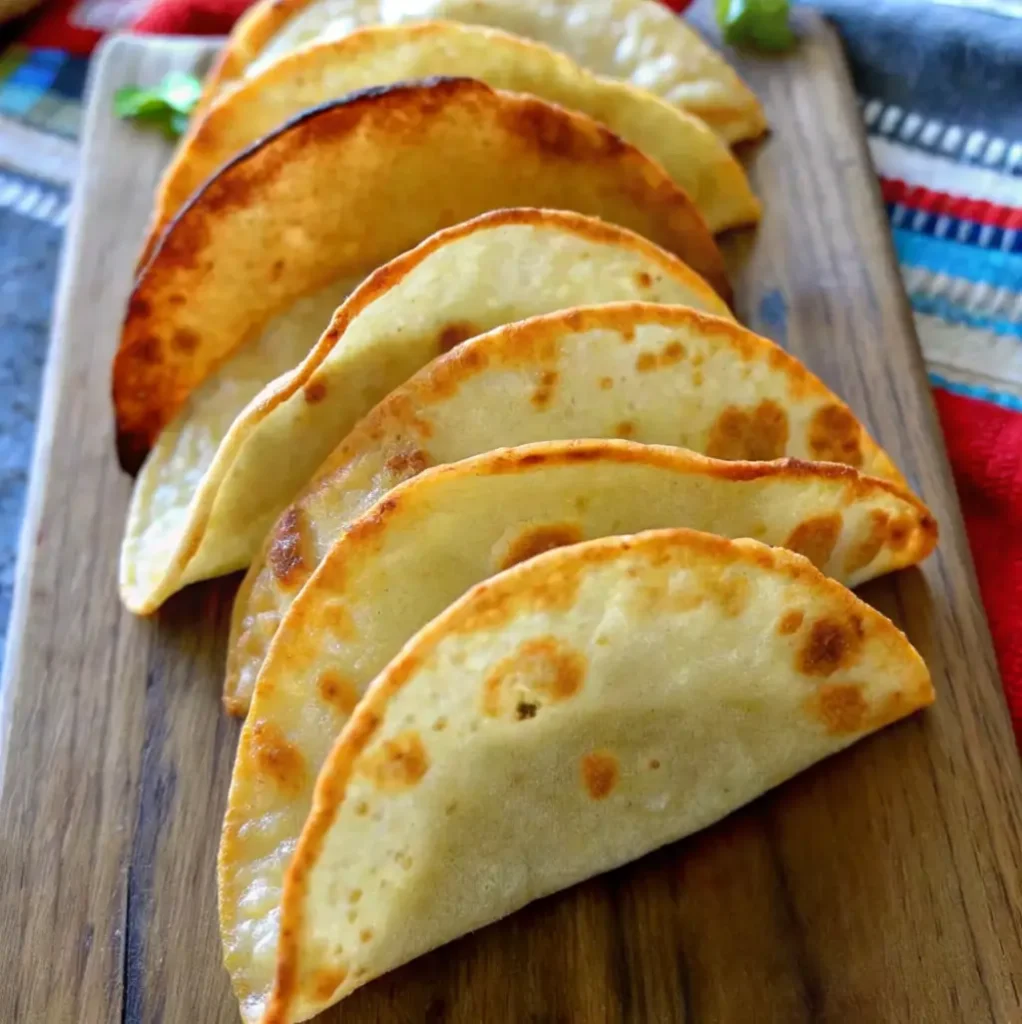 A high-angle view of a row of crispy, folded taco shells (likely corn or flour) with golden-brown, slightly fried edges, arranged diagonally on a wooden cutting board.