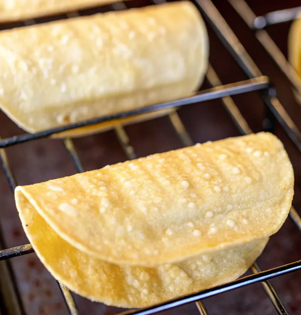 Several yellow corn tortilla shells are folded and heated on a wire baking rack, showing a slightly crispy and blistered texture, ready to be filled for tacos.