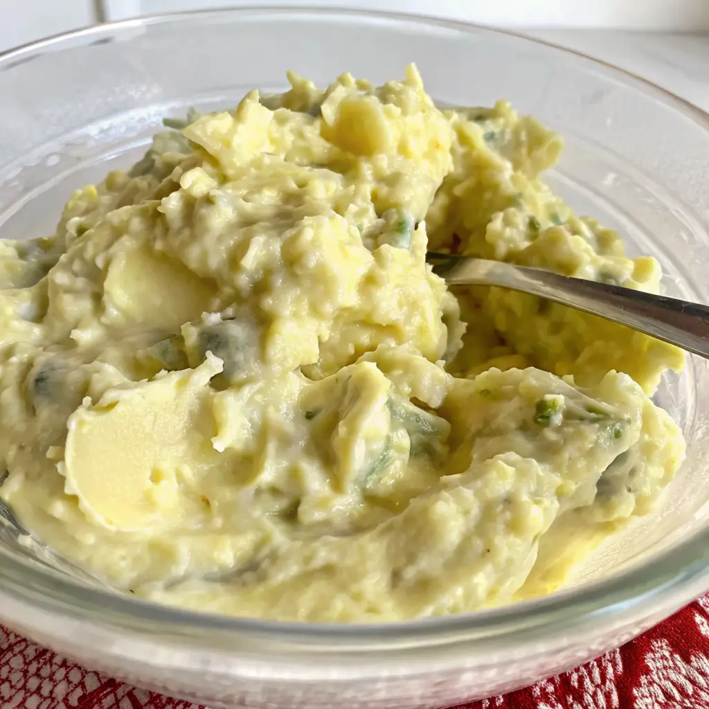 A clear glass bowl holds a creamy, pale yellow mashed potato salad, generously mixed with bright green herbs like parsley or chives.