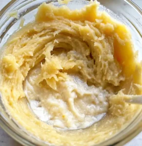 A close-up of the mashed flesh of the roasted squash being stirred in a clear glass bowl, showing a thick, creamy, pale-yellow mixture, likely with added butter or cream.