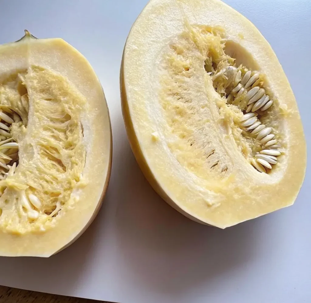 Two halves of a raw Mashed Potato Squash (Sweet Dumpling Squash) are cut open, revealing the stringy, pale-yellow flesh and the cluster of seeds in the center cavity.