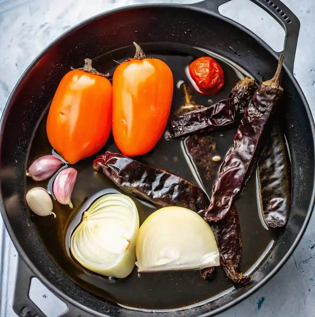 Fresh vegetables including tomatoes, onion halves, and garlic cloves are simmering in a dark cast-iron pan with dried chiles, water, as part of the Mexican hot sauce preparation.