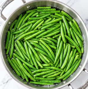 Fresh whole green beans resting in a stainless steel pot filled with water, viewed from directly above on a marble surface.