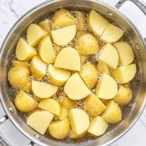 Yellow potatoes, cut into quarters, are simmered in salted water in a stainless-steel pot on a marble countertop.