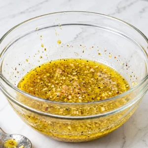 Homemade vinaigrette, mixed with minced garlic, mustard, black pepper, olive oil, and lemon in a clear glass mixing bowl on a marble countertop.
