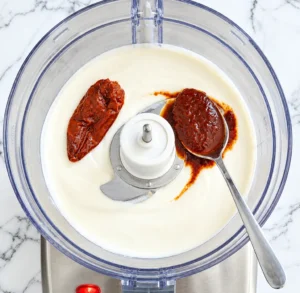 Top view of a food processor filled with a base of white ranch dressing, topped with a whole chipotle pepper and a spoonful of spicy adobo sauce.