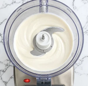 A food processor containing mayonnaise, sour cream, and buttermilk, ready to be mixed on a marble countertop.