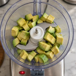 Fresh avocado cubes arranged in the bowl of a clear food processor, ready to be blended into a homemade ranch dressing.