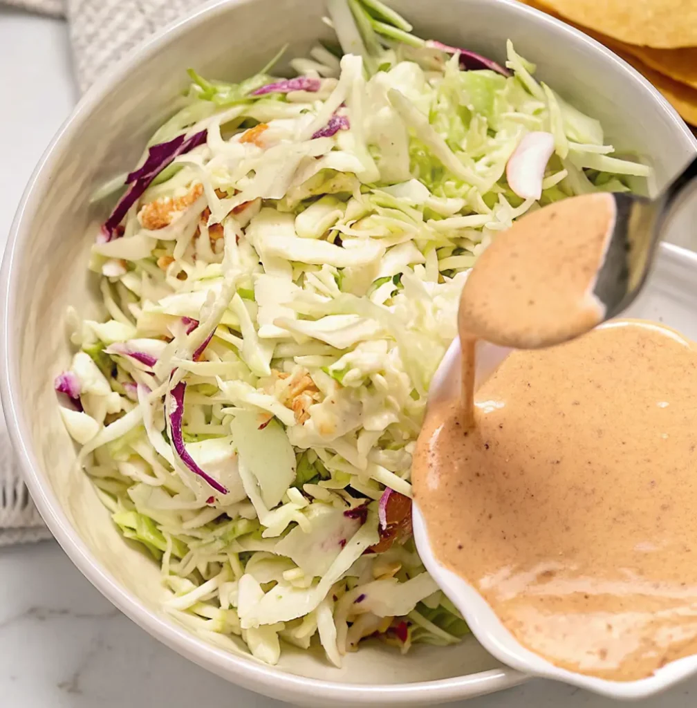 A white bowl of shredded cabbage and radicchio slaw is being topped with a spoonful of creamy Baja taco sauce. The dressing is a light orange-brown color with visible specks of spice.