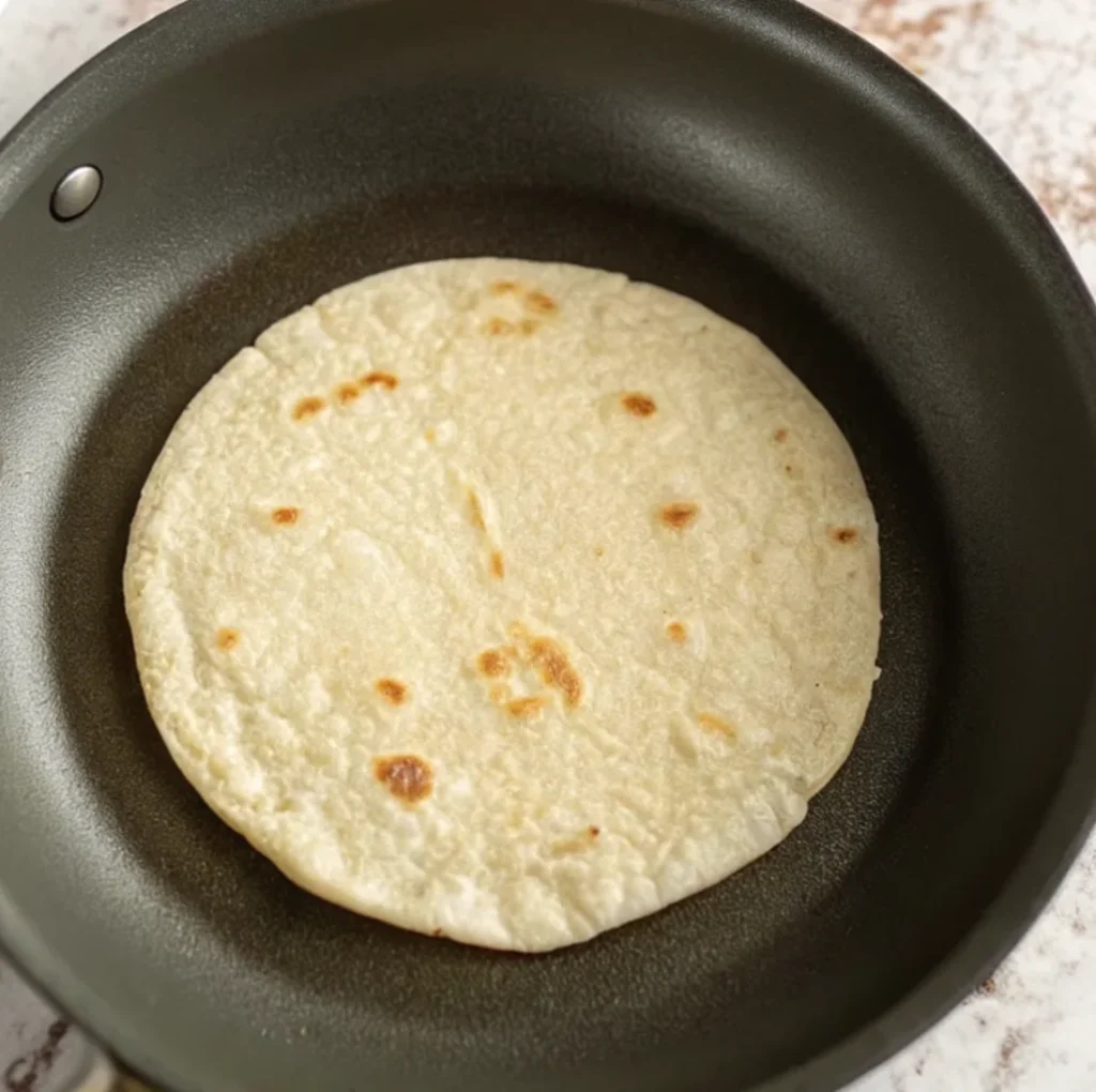 A single white corn or flour tortilla is warming up in a black non-stick skillet. It has developed a few lightly browned, toasted spots from the heat, perfect for making tacos.