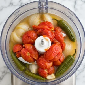 Top view of a food processor containing cooked tomatoes, whole green jalapeño peppers, onions, and broth, ready to be blended into a Mexican sauce.