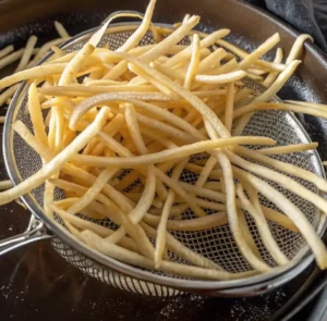 golden-yellow shoestring potato fries draining in a metal mesh strainer, suggesting they were freshly fried and are crispy and salty.