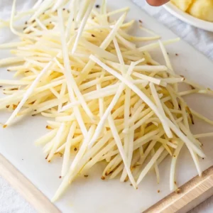 pile of raw, thin shoestring potato strips on a white cutting board with a wooden edge. The strips have bits of potato skin remaining, indicating they were freshly cut.