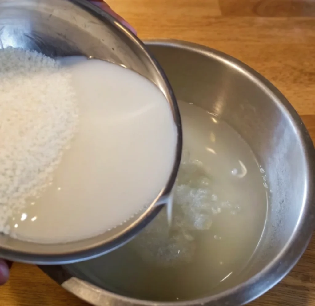 A bowl of uncooked white rice soaked in water filled with starch is drained into a larger metal bowl.