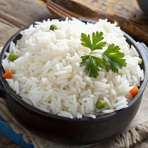 Steam cooked rice recipe is served in a black decorative bowl on a wooden table, garnished with a sprig of fresh parsley and topped with small pieces of carrots and green peas.
