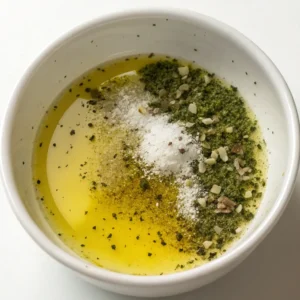 Close-up of a small white bowl holding olive oil, lemon juice, oregano, salt, and pepper to make a simple Mediterranean salad dressing.