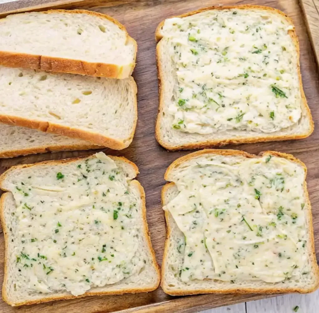 four slices of Texas Toast on a wooden tray. The two slices on the right are generously spread with the light green and creamy garlic herb butter mixture, while the two slices on the left remain plain.