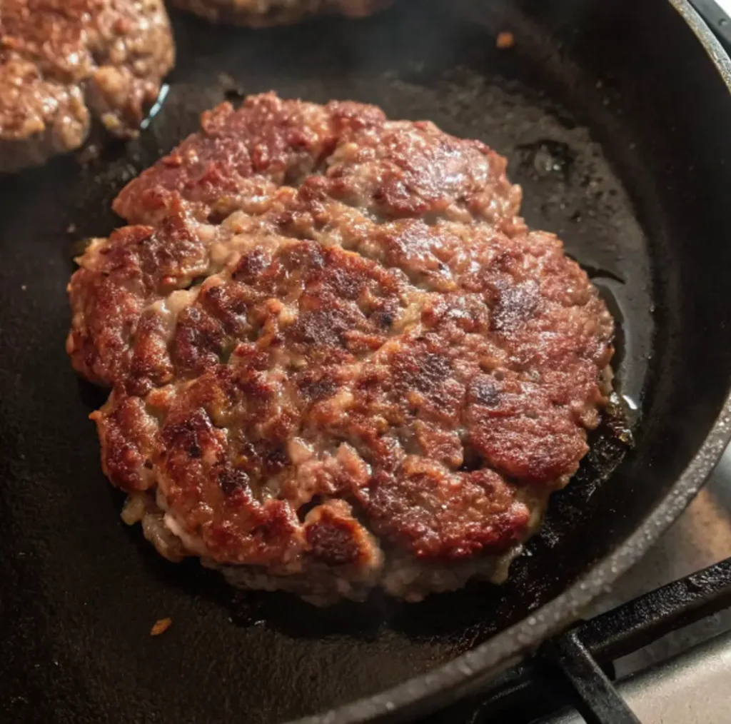 Wagyu beef patty is shown in a hot cast-iron skillet, developing a deep brown crust as it cooks to perfection, promising a rich, savory burger.