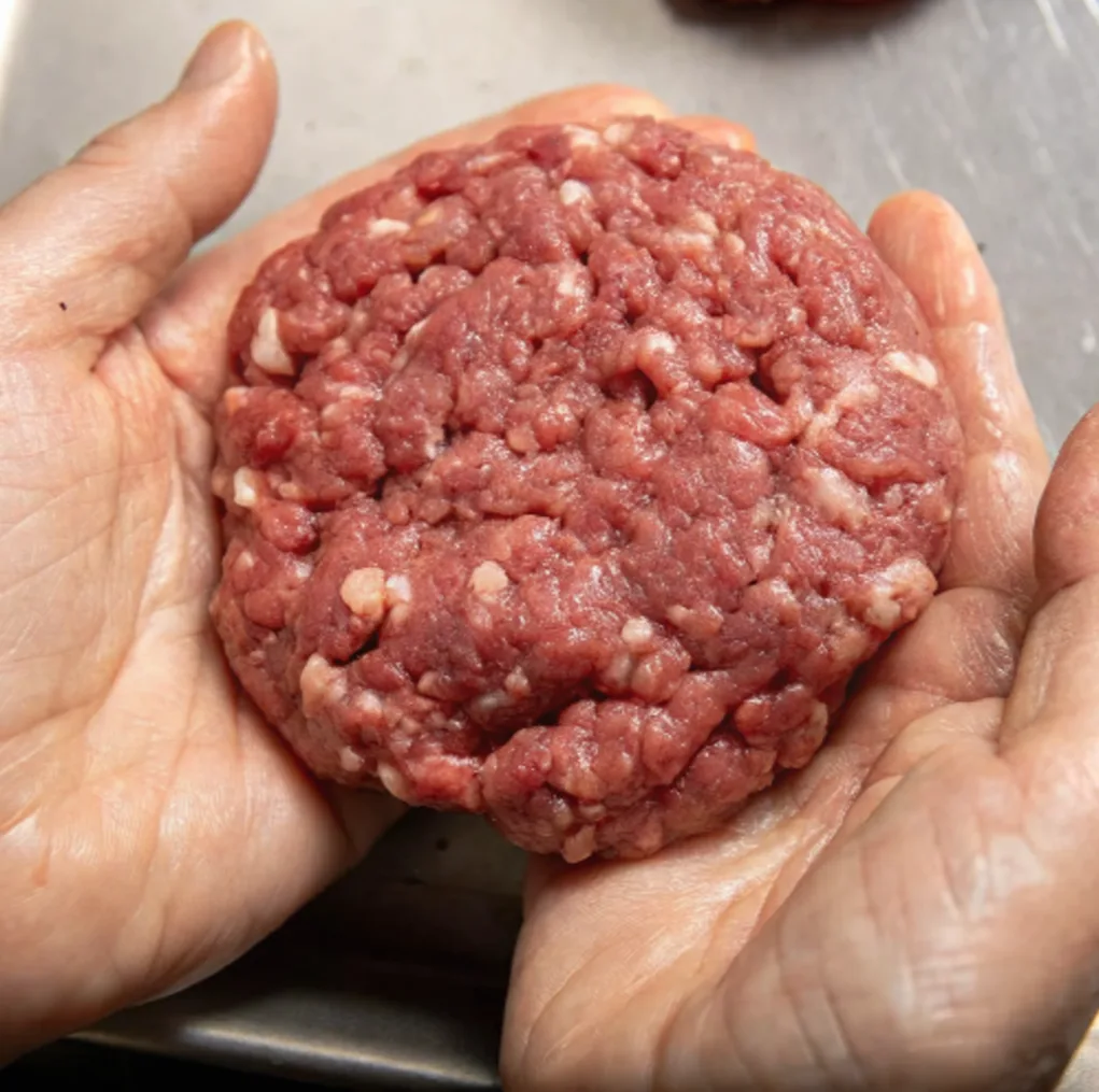 A person's hands gently cup a raw, circular patty made from ground Wagyu beef. The meat has a rich red color speckled with abundant white fat