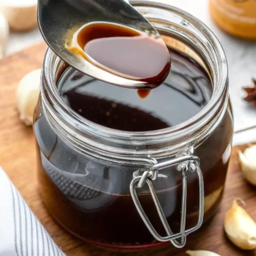 A spoonful of smooth, rich, and dark brown Worcestershire sauce is lifted from a glass jar, showcasing its glossy texture.