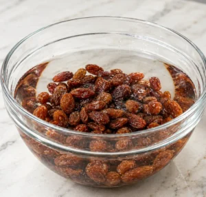Dark plump raisins soaking in water in a clear glass bowl on a marble surface for homemade Worcestershire sauce preparation.