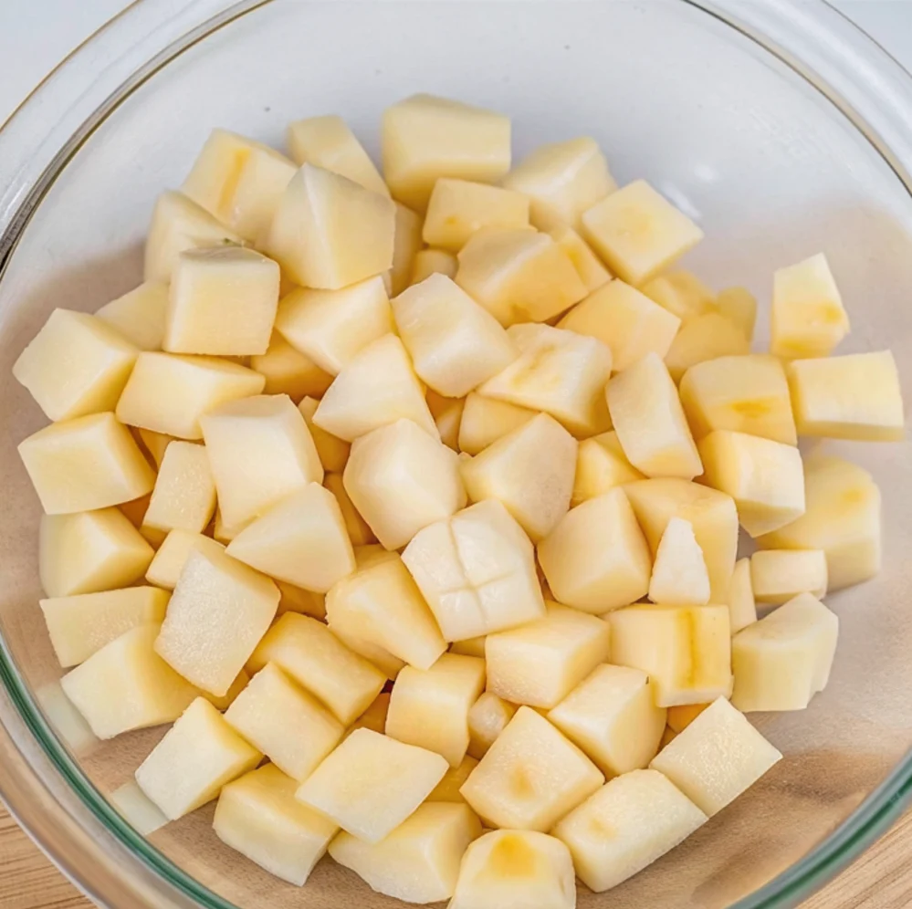 A clear glass bowl is filled to the brim with peeled and uniformly diced raw potatoes. These little cubes are prepped