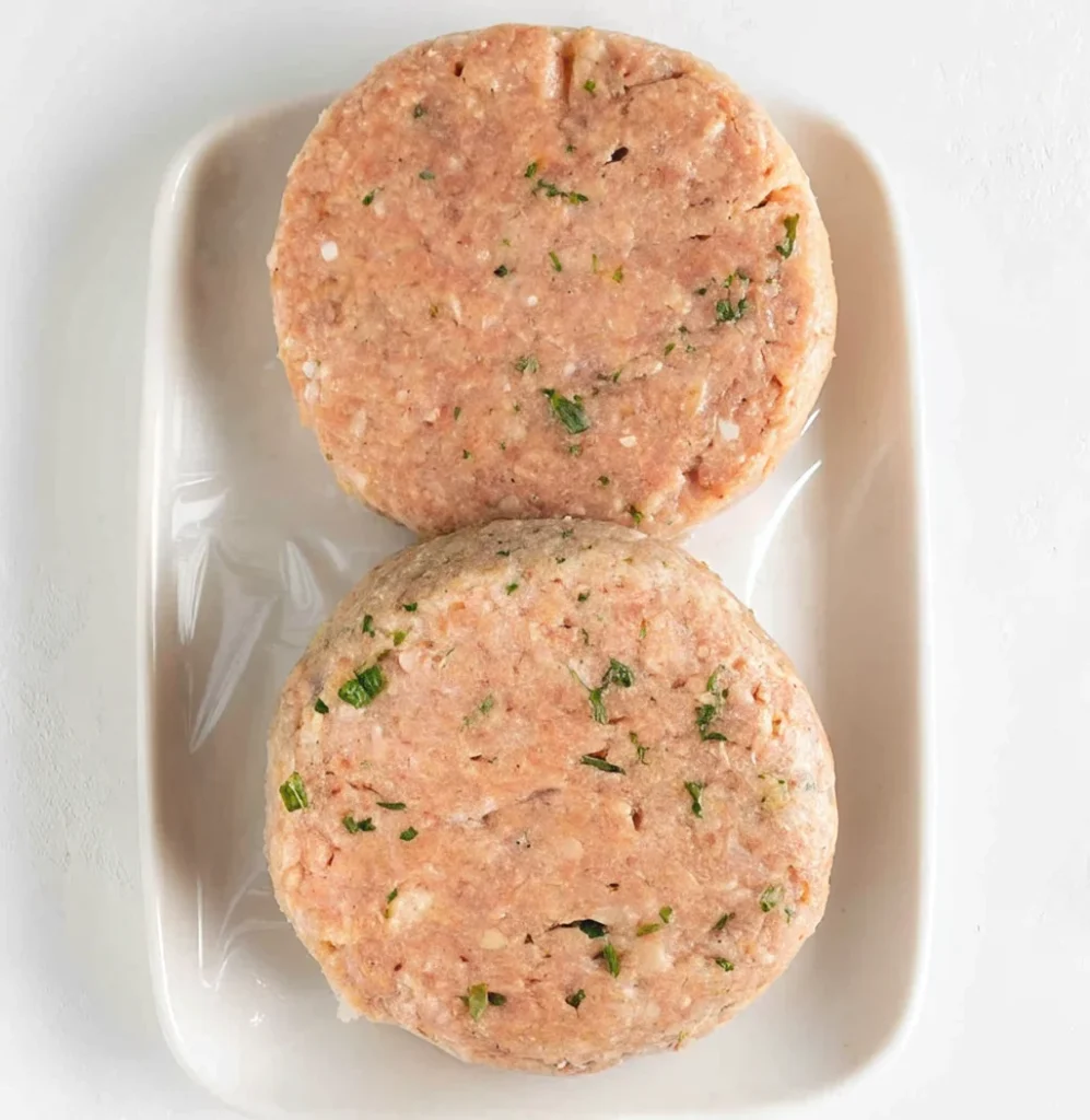 Two raw, seasoned ground turkey burger patties with visible flecks of green herbs are placed on a white rectangular plate, ready to be cooked in an air fryer.