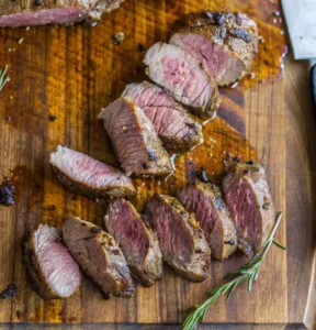 Succulent slices of seared lamb tenderloin are fanned out on a wooden cutting board, showcasing a juicy pink center, a dark herb-crusted exterior, and a garnish of fresh rosemary.