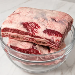 Thickly cut raw beef plate ribs with heavy fat caps and rich red marbling, stacked inside a clear glass bowl resting on a white marble surface.