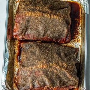Two thick slabs of seasoned raw beef plate ribs with a dark spice rub, resting in a foil-lined baking pan with flavorful juices pooling around the meat.