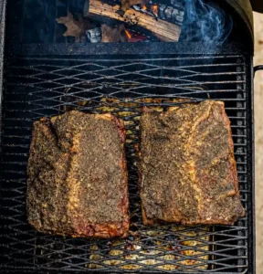 Two seasoned beef plate ribs cooking side by side on a smoker rack, positioned directly above burning wood logs generating subtle wisps of smoke.