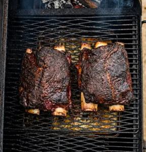Two racks of fully smoked beef plate ribs with a rich, dark caramelized bark resting on a grill grate inside an active smoker setup.