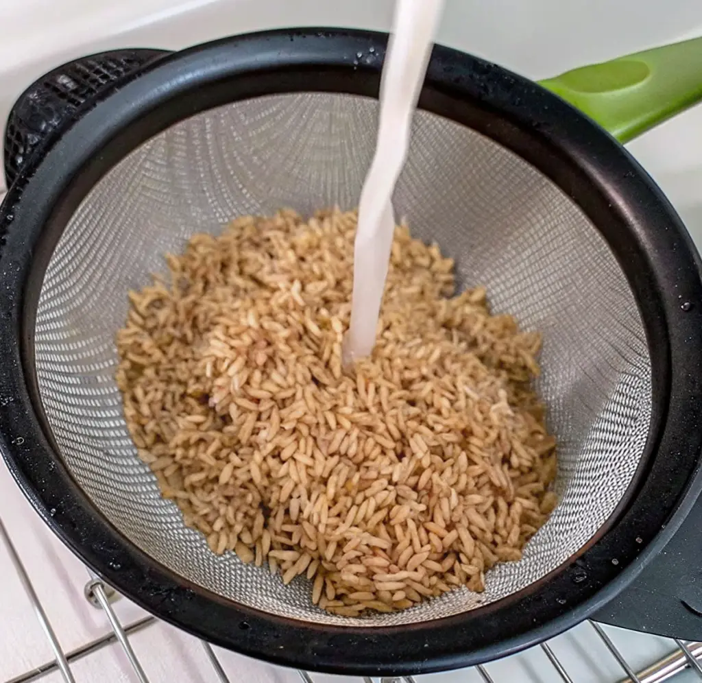 A stream of water rinses uncooked brown rice in a fine-mesh strainer, washing away excess starch to ensure fluffy, non-sticky results before cooking in an Instant Pot.
