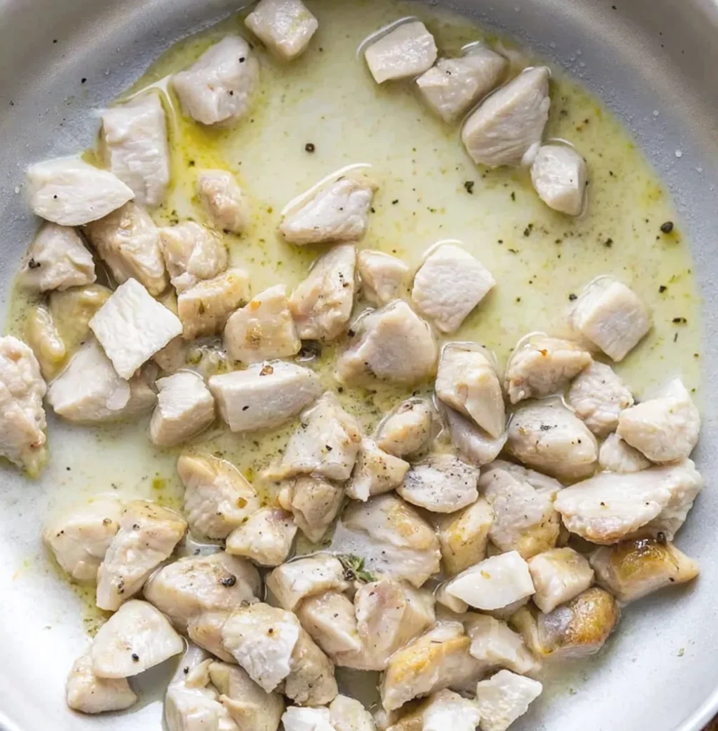 Small pieces of chicken are being seared in a silver pan with oil and black pepper. The chicken is turning golden brown as it cooks in the shimmering liquid.
