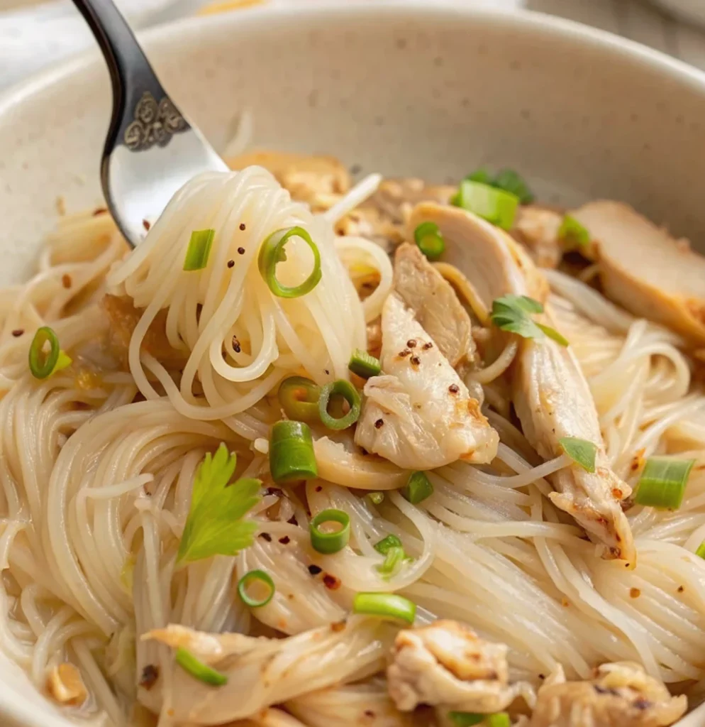 A close-up of chicken long rice recipe featuring clear bean thread noodles, tender shredded chicken, and sliced green onions in a savory broth, served in a speckled ceramic bowl.
