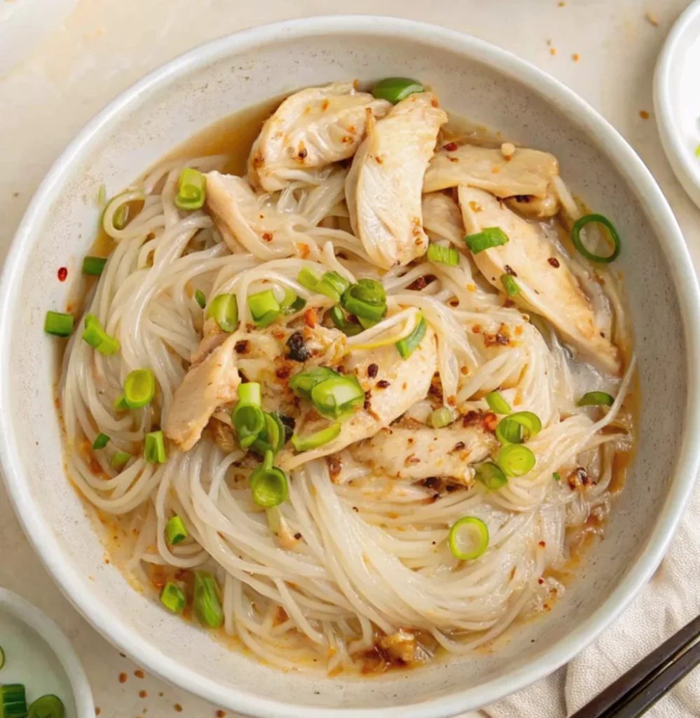 A close-up of chicken long rice recipe featuring clear bean thread noodles, tender shredded chicken, and sliced green onions in a savory broth, served in a speckled ceramic bowl.
