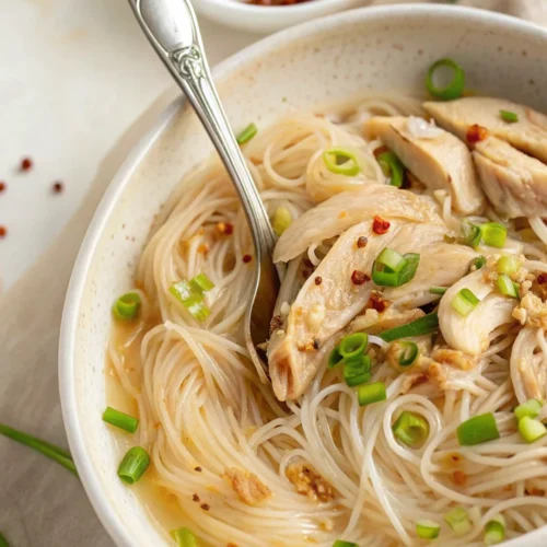 A close-up of chicken long rice recipe featuring clear bean thread noodles, tender shredded chicken, and sliced green onions in a savory broth, served in a speckled ceramic bowl.