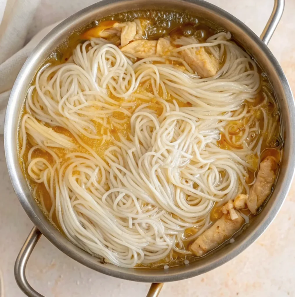 The softened long rice noodles and shredded chicken combined in the pot, simmering together in the golden broth until the noodles absorb the savory ginger and soy flavors.